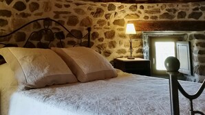 A bedroom with a stone wall, a bed with pillows, a nightstand with a lamp, and a window with a view. - Windmill gite unusual (Auvergne)
