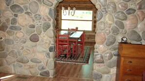 A stone-walled dining area with a wooden table and chairs, a window with curtains, and a clock on a wooden dresser.
