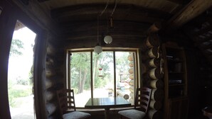 A log cabin interior with a wooden dining table, two chairs, a hanging light fixture, and a large window offering a view of the outdoors.