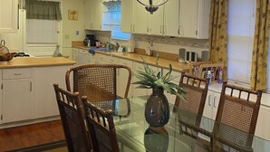 Un comedor con una mesa de vidrio rodeada de sillas de madera, un candelabro y una cocina al fondo.