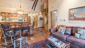 A spacious living area with a high ceiling, a chandelier, a leather sofa with decorative pillows, a wooden dining table with chairs, and a kitchen in the background. - 3/2 Ski In/Out  BEAVER CREEK, steps to Village, Townsend Place A307 (Beaver Creek)