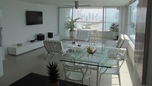 A modern living room with a glass dining table, white chairs, a mounted TV, and a balcony with a city view. - Oceanfront Penthouse with Stunning Views of the Caribbean. (Cartagena)