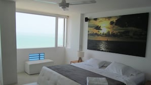 A modern bedroom with a large bed, a bench by the window, and a painting of a sunset over water. - Oceanfront Penthouse with Stunning Views of the Caribbean. (Cartagena)