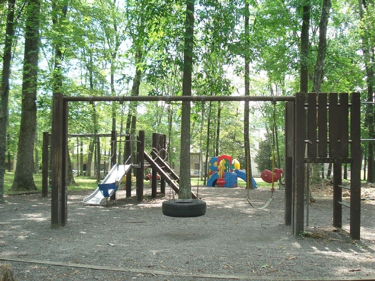 Un parque infantil con un columpio de neumático, un tobogán y una estructura para escalar en un área boscosa.