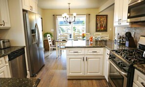 Une cuisine avec des meubles blancs, un réfrigérateur en acier inoxydable, une cuisinière à gaz et un coin repas agrémenté d’un lustre.