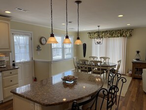 A kitchen with a granite island, pendant lights, a dining area with a table and chairs, and a window with sheer curtains.