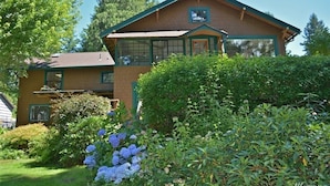 A two-story house with a brick exterior and green trim, surrounded by lush greenery and hydrangeas. - PEACEFUL WATERFRONT RETREAT 30-MINUTE FERRY RIDE FROM SEATTLE! (Bremerton)