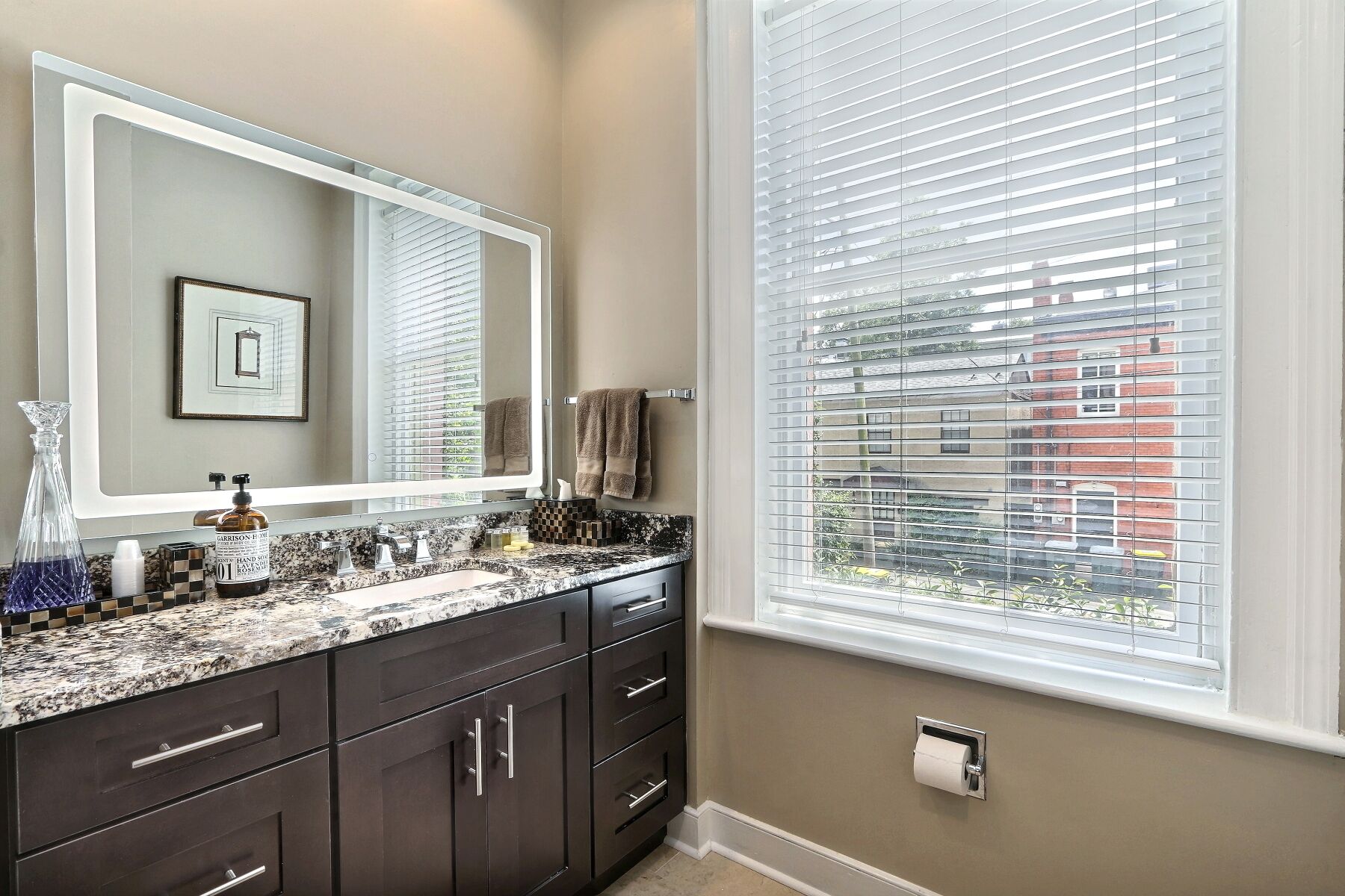 A bathroom with a large mirror, dark cabinets, a granite countertop, and a window with blinds.