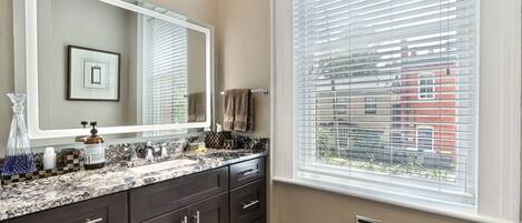 A bathroom with a large mirror, dark cabinets, a granite countertop, and a window with blinds.