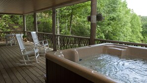 Un bain à remous sur une terrasse en bois, avec des fauteuils à bascule blancs et une vue sur une forêt.