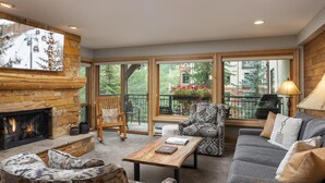 A cozy living room with a fireplace, comfortable seating, and large windows offering a view of the outdoors.