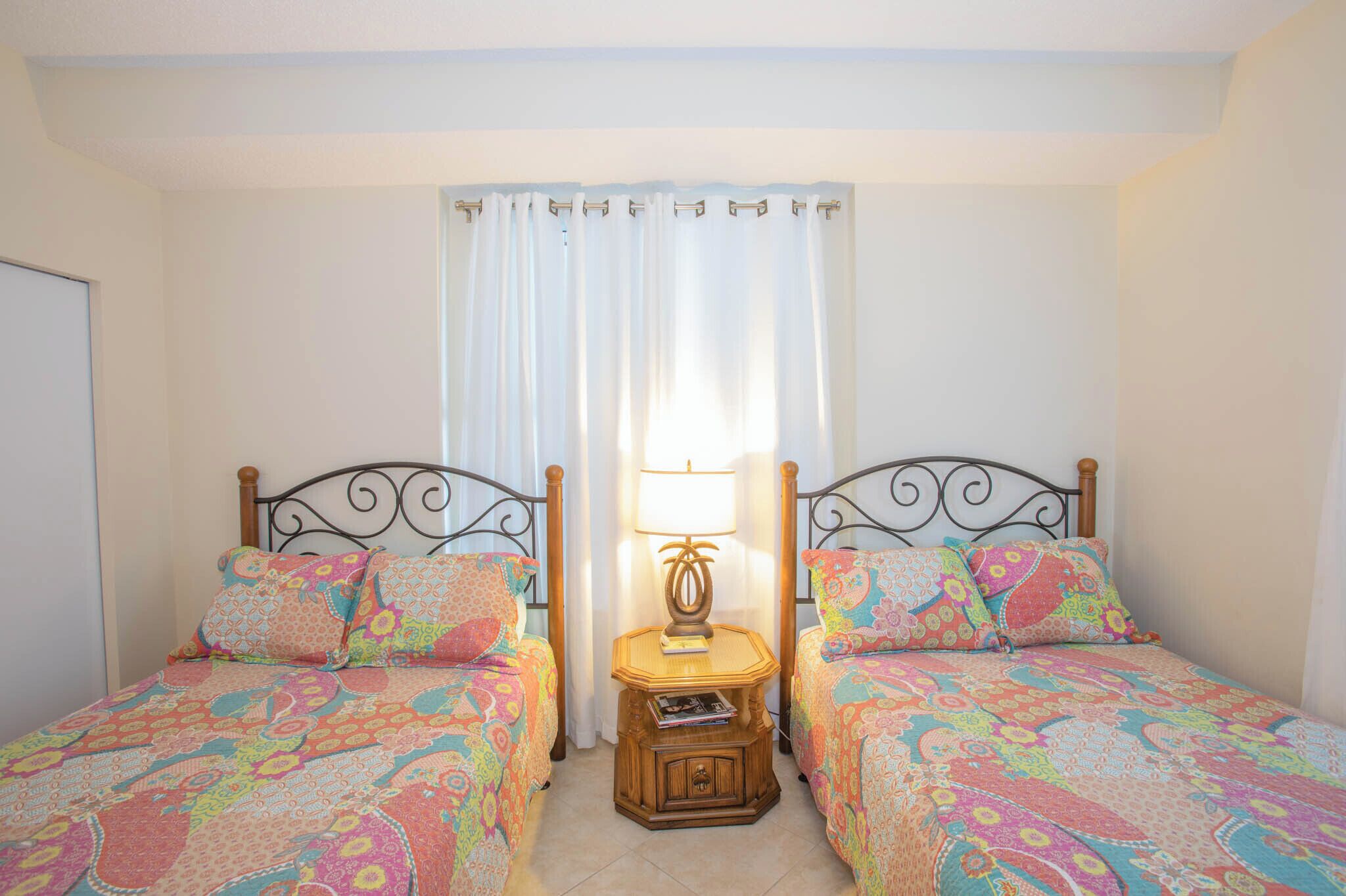 A bedroom with two beds, each with patterned bedding, a bedside table with a lamp, and a window with curtains.