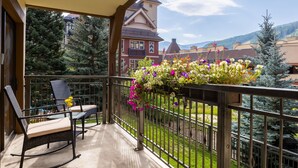A balcony with a rocking chair and a table, overlooking a garden with flowers and a view of buildings and trees. - Montaneros 218 -2 Bedroom Diamond (Vail)