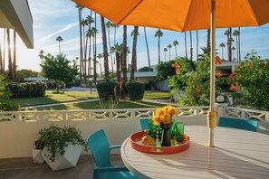 A patio with a round table, chairs, and an umbrella. There are potted plants and a view of palm trees and a lawn. - Mid-Century Luxury Condo near Downtown Palm Springs (Palm Springs)