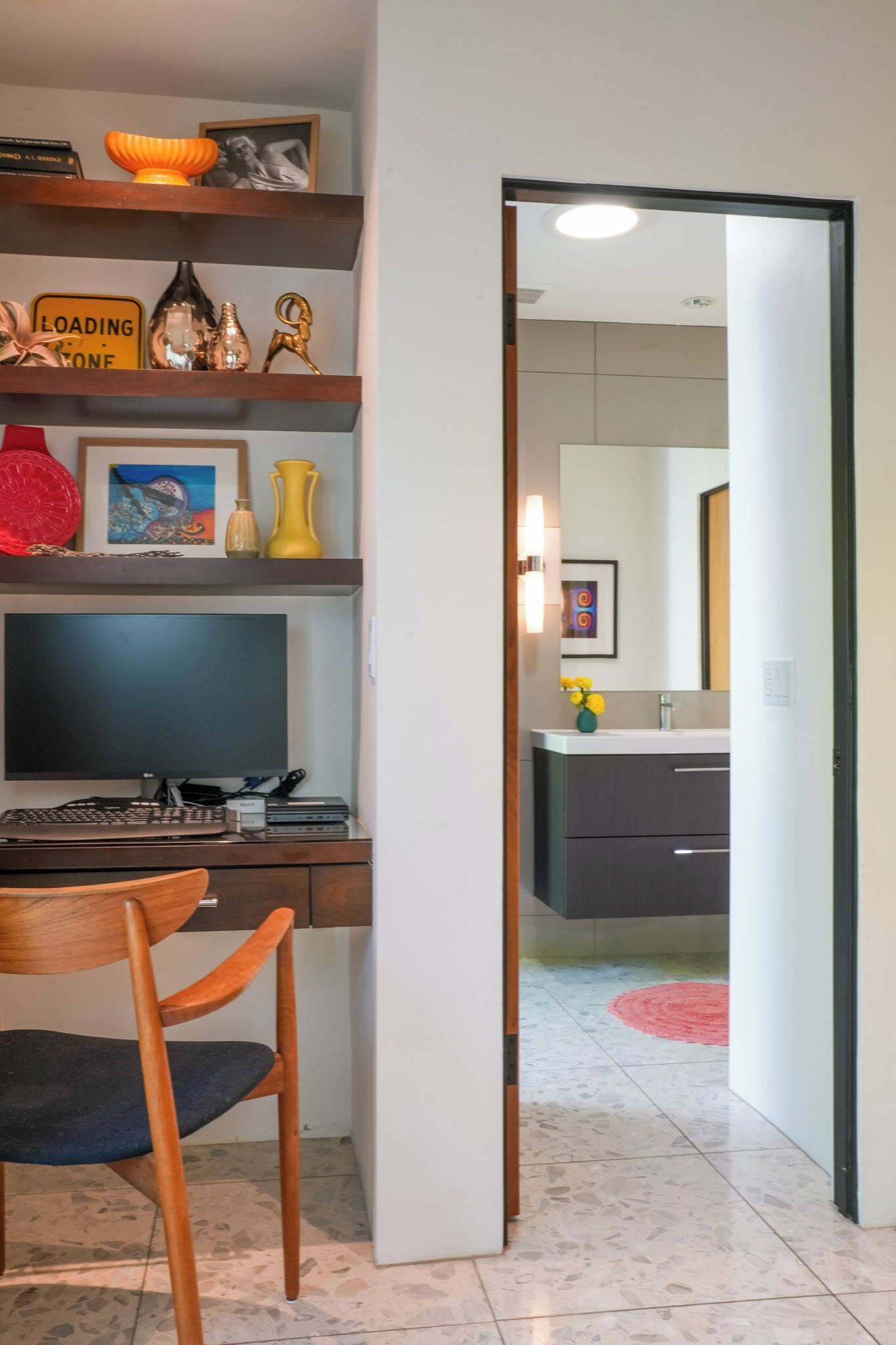 Ein Arbeitszimmer mit Schreibtisch, Computer und Stuhl. Eine Tür führt zu einem Badezimmer mit Waschbecken und Spiegel.