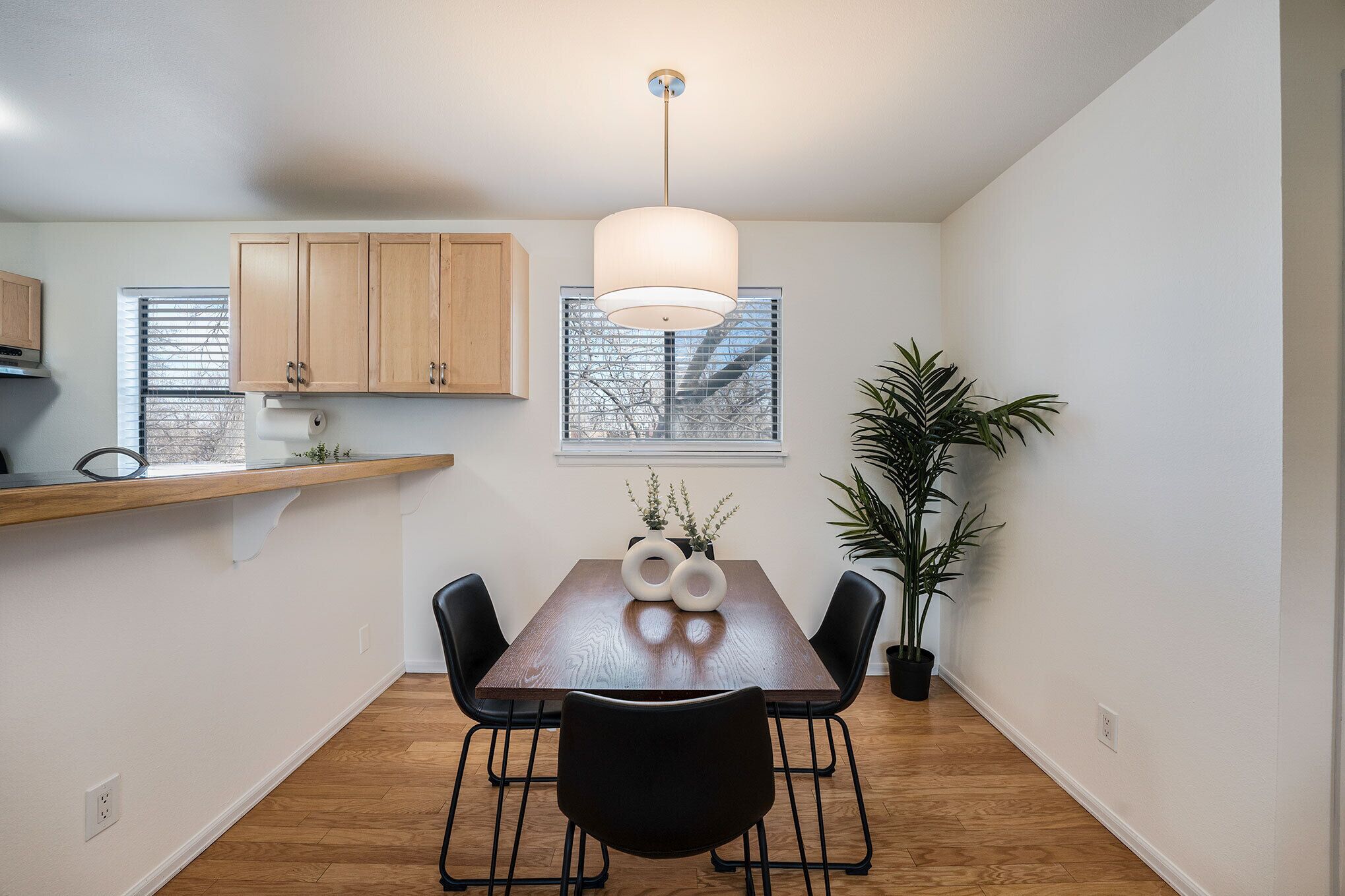 A dining area with a wooden table, black chairs, a pendant light, and a potted plant.