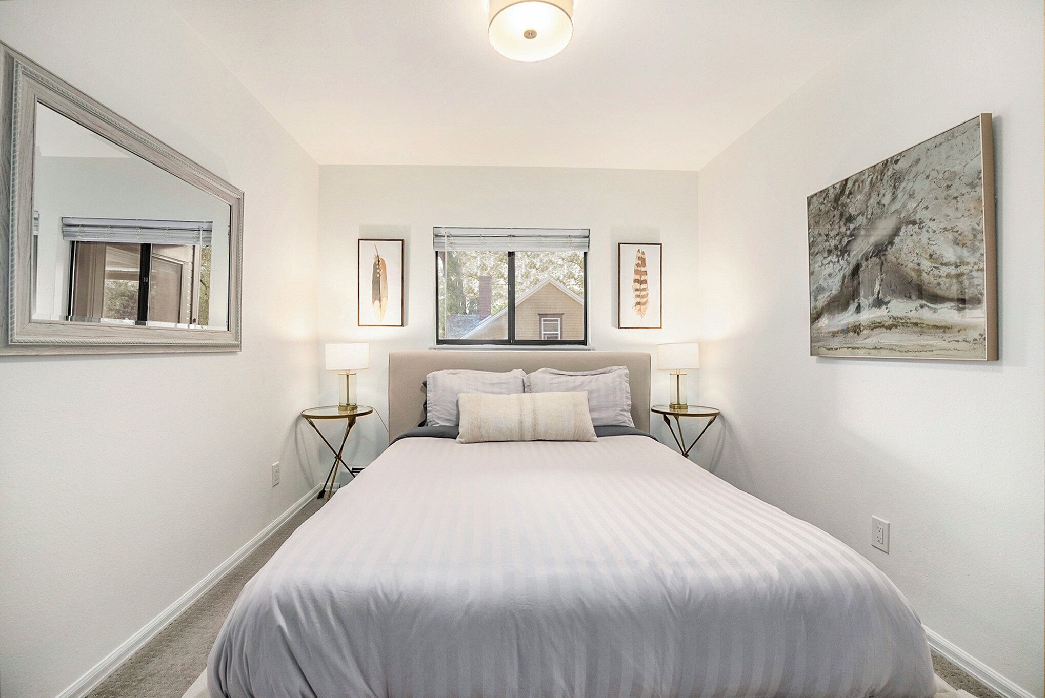 A bedroom with a large bed, two bedside tables with lamps, a window with blinds, and framed pictures on the walls.