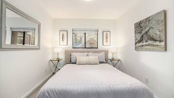A bedroom with a large bed, two bedside tables with lamps, a window with blinds, and framed pictures on the walls.