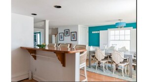Una cucina moderna con isola in legno, mobili bianchi e zona pranzo con sedie bianche e una parete azzurra a contrasto.