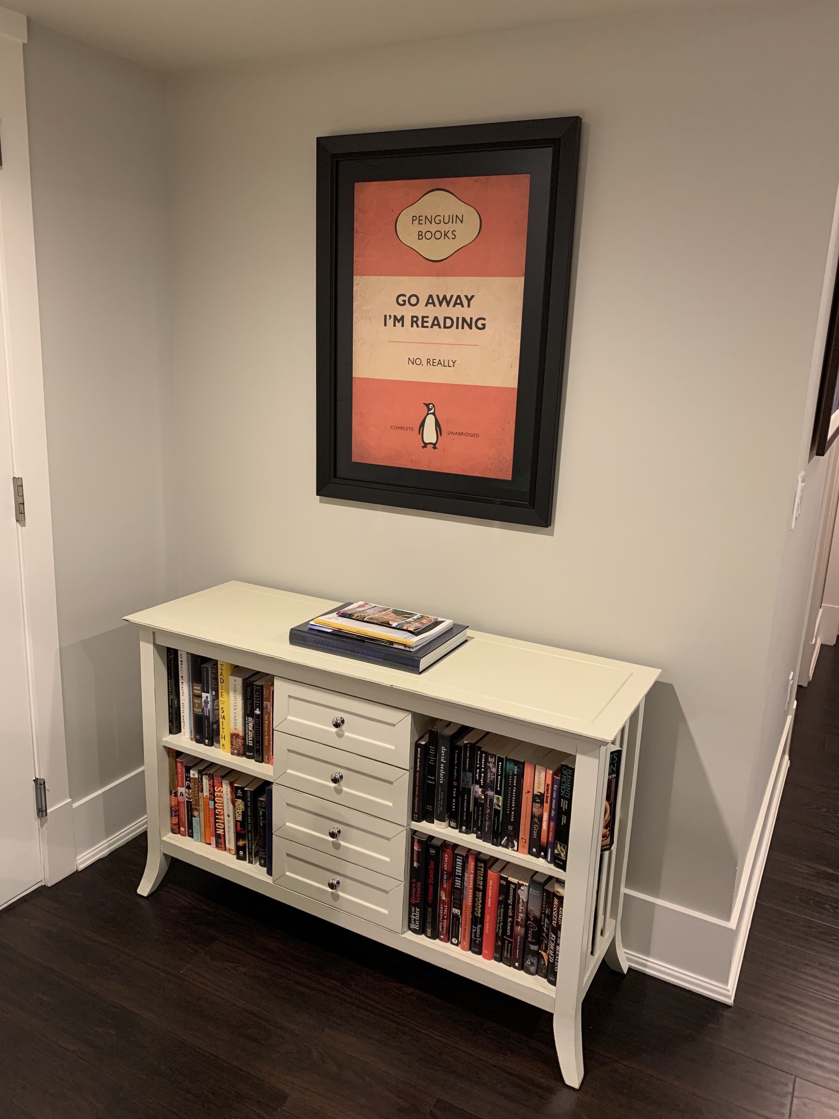 A white bookshelf with multiple shelves, a framed poster on the wall above it, and a door to the left.