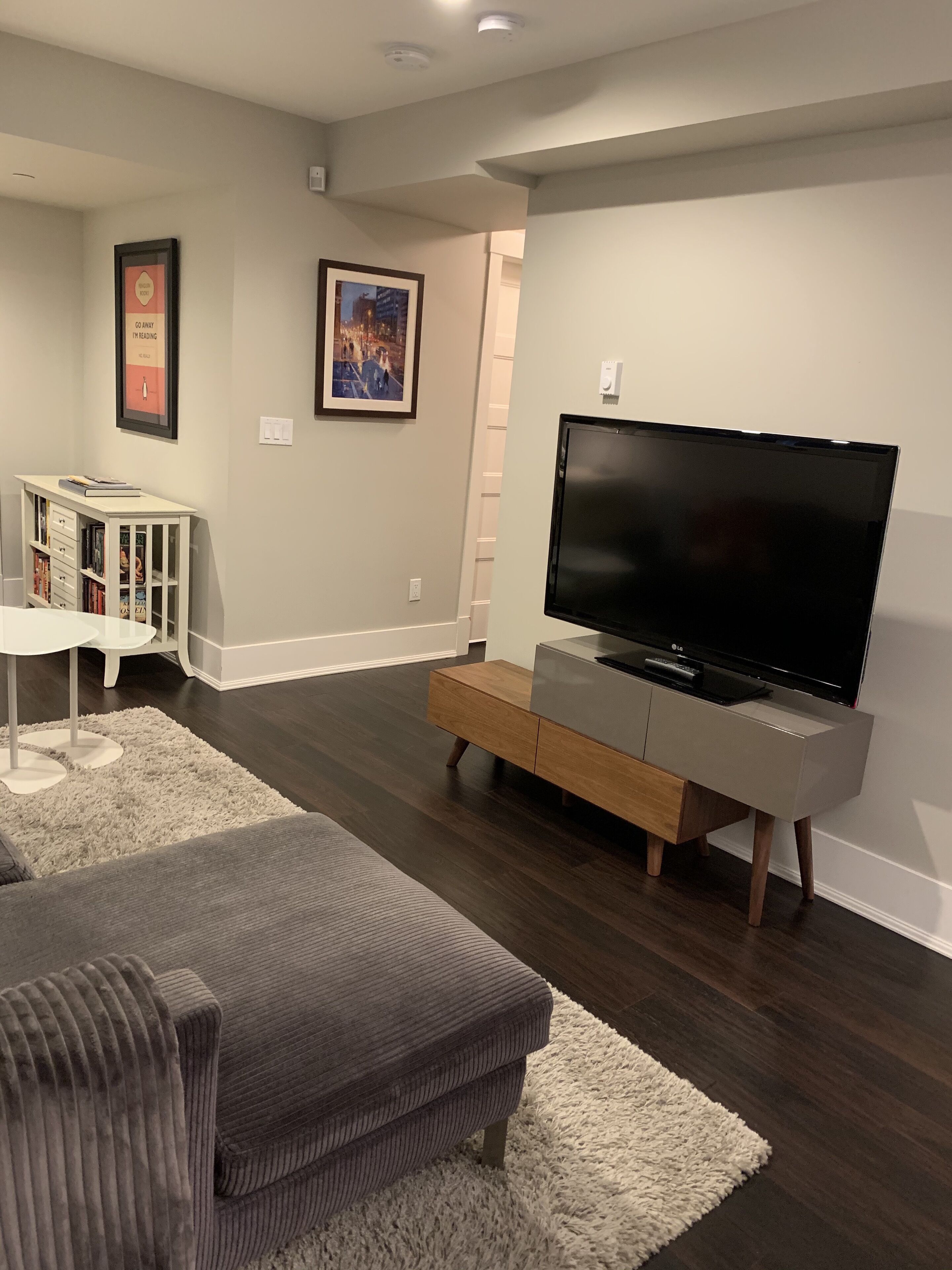 A living room with a flat-screen TV, a white entertainment unit, a gray sofa, a white rug, and a bookshelf.