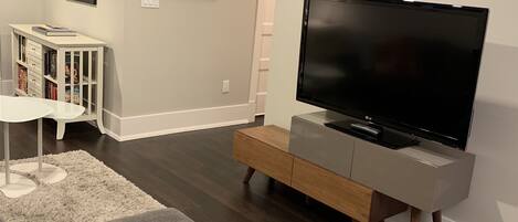 A living room with a flat-screen TV, a white entertainment unit, a gray sofa, a white rug, and a bookshelf.