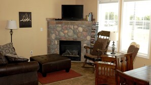 Una sala de estar con chimenea de piedra, televisor montado arriba y un área de descanso con sofá, reposapiés y sillones.