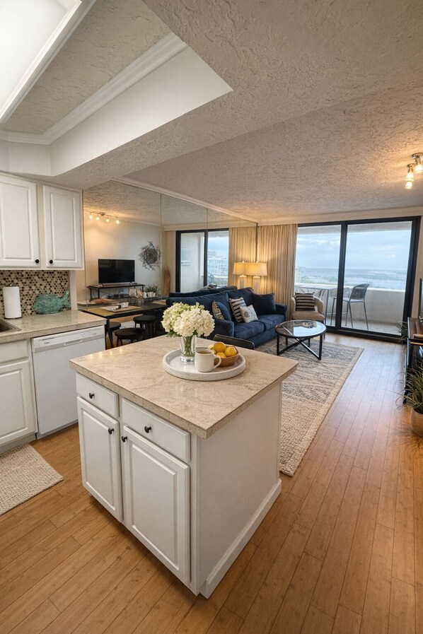A modern kitchen with a central island, white cabinetry, and a dining area with a blue sofa and a view of the ocean.