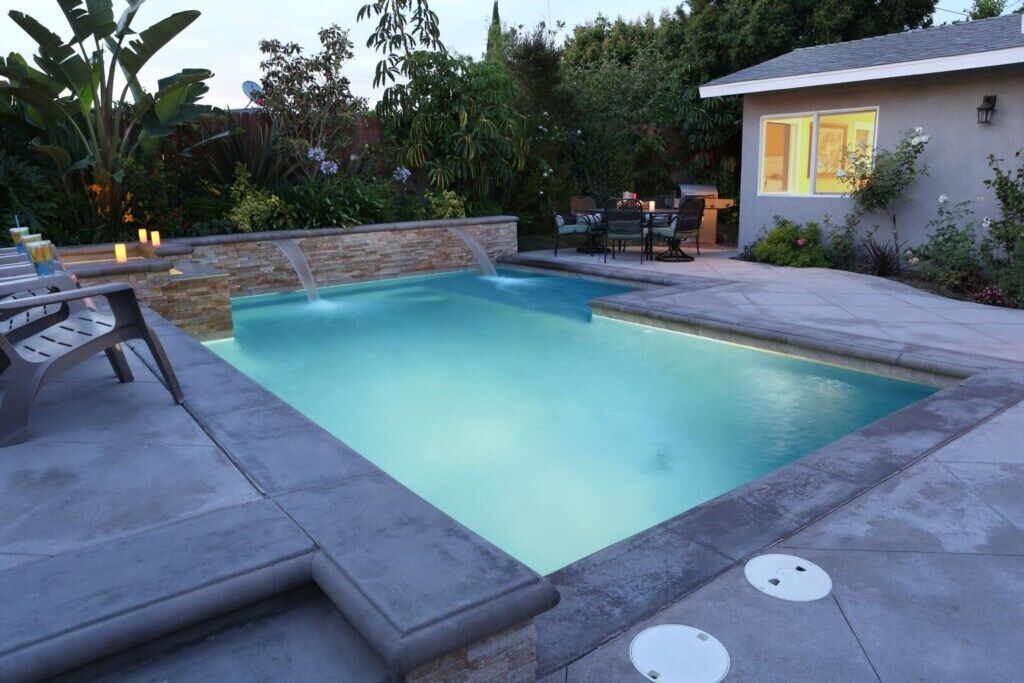 Une piscine rectangulaire bordée de pierres, un espace patio avec des meubles d’extérieur, et une maison en arrière-plan.