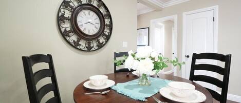 A dining area with a round wooden table, two chairs, a wall-mounted clock, and a vase with flowers.