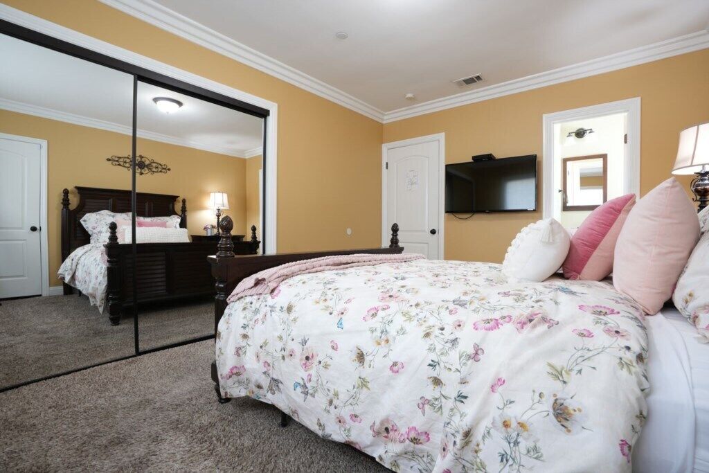 A bedroom with a large mirror, a bed with floral bedding, a television on the wall, and a door with a light switch.