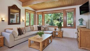 Ein gemütliches Wohnzimmer mit Holzboden, großen Fenstern, einer Couch, einem Couchtisch und einem Fernseher an der Wand.