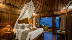 A wooden room with a bed, a nightstand, a table, and a view of the outside. - Mandana Ubud Villa (Ubud)