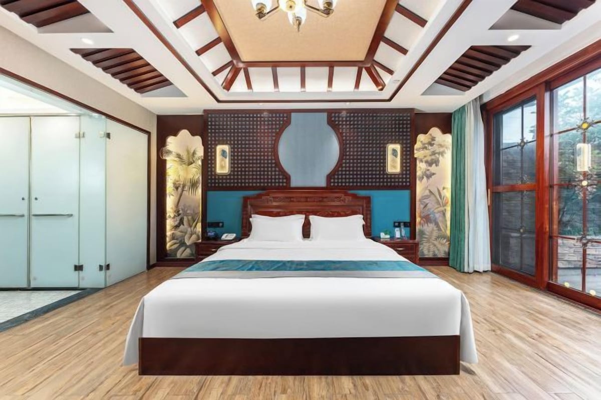 A spacious bedroom with a large bed, wooden ceiling beams, and a large window offering a view of the outdoors.