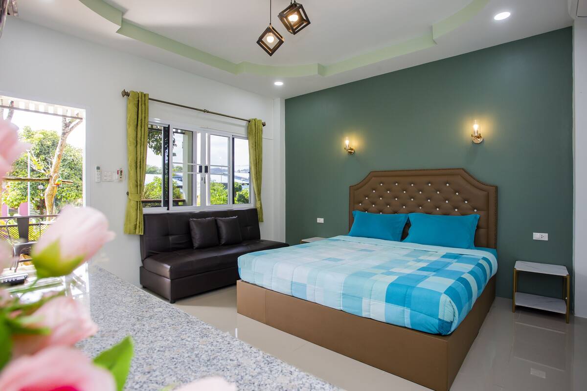 Ein Schlafzimmer mit Bett, einer Couch, einem Fenster mit Vorhängen und Blick auf Grünfläche.