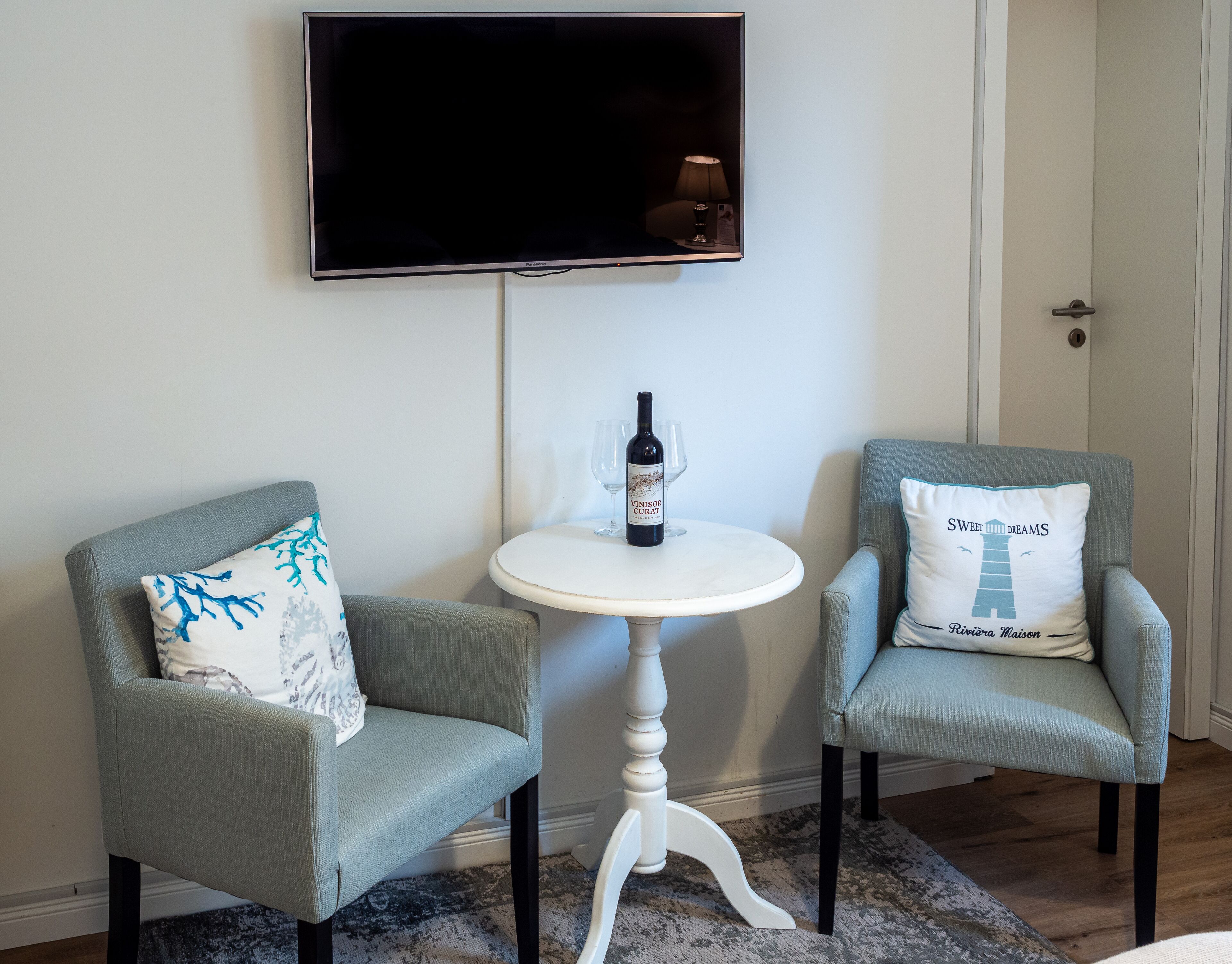 Une pièce comprenant deux fauteuils, une petite table ronde avec une bouteille de vin et des verres, et un téléviseur à écran plat fixé au mur.