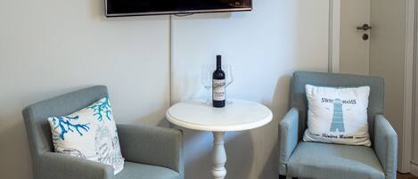 Une pièce comprenant deux fauteuils, une petite table ronde avec une bouteille de vin et des verres, et un téléviseur à écran plat fixé au mur.