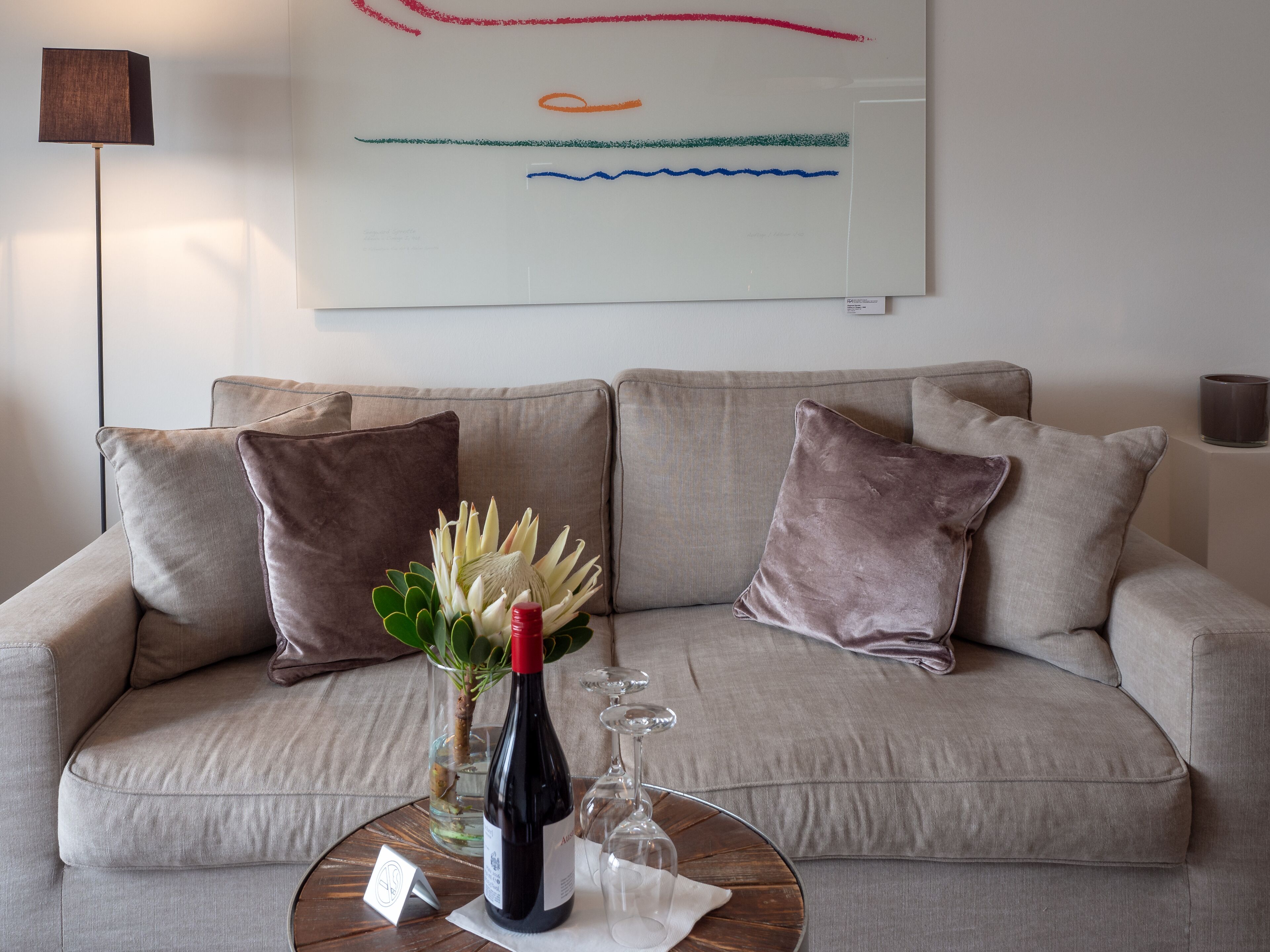 A living room with a beige sofa, two brown cushions, a glass vase with flowers, a wine bottle, and a candle on a wooden coffee table.