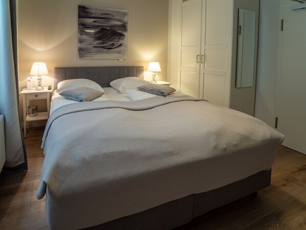 A neatly made bed with a textured blanket, two bedside lamps, a wall-mounted artwork, and a closed door. - Boutique Hotel Villa Monika (Sylt)