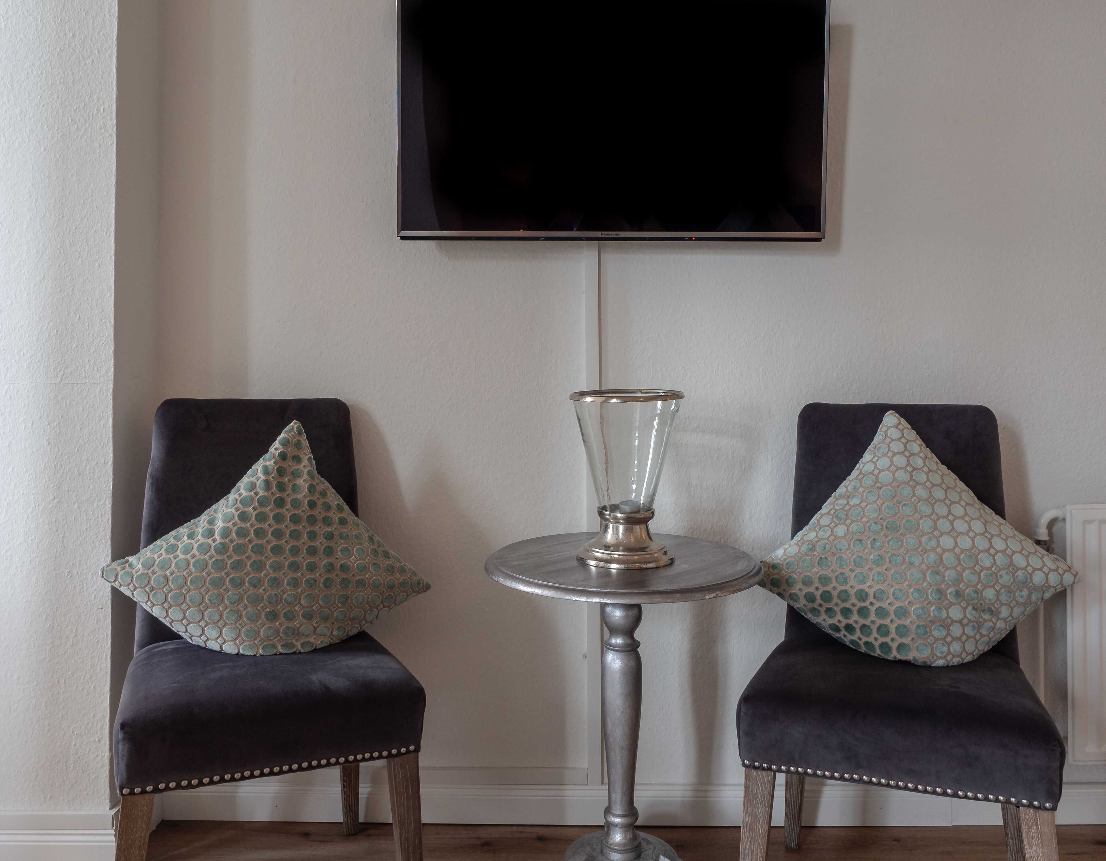 Two dark upholstered chairs with patterned cushions, a small round table with a glass object, and a flat-screen TV mounted on the wall.