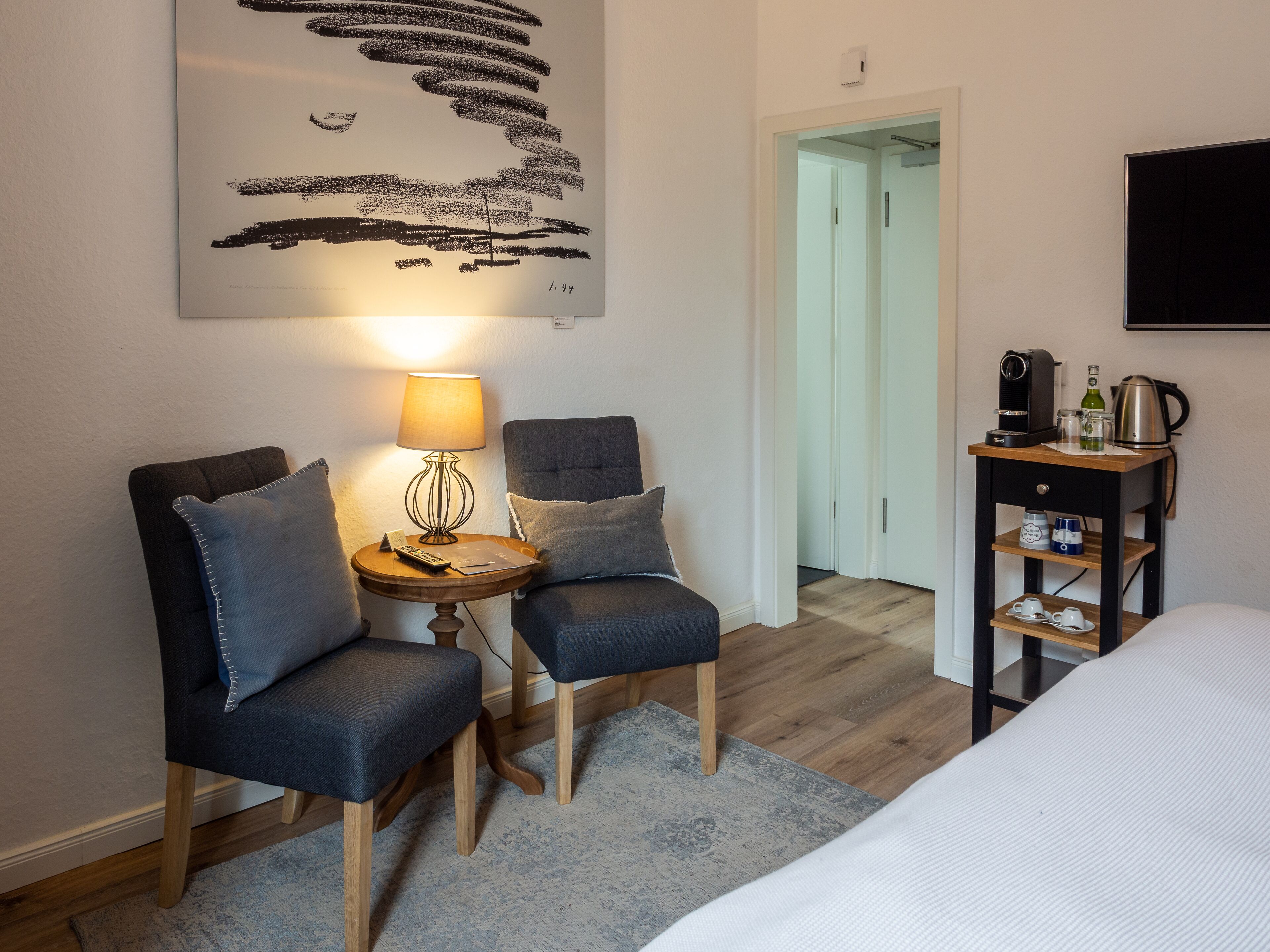 A hotel room with two armchairs, a small table with a lamp, a bed, a coffee station, and a painting on the wall.