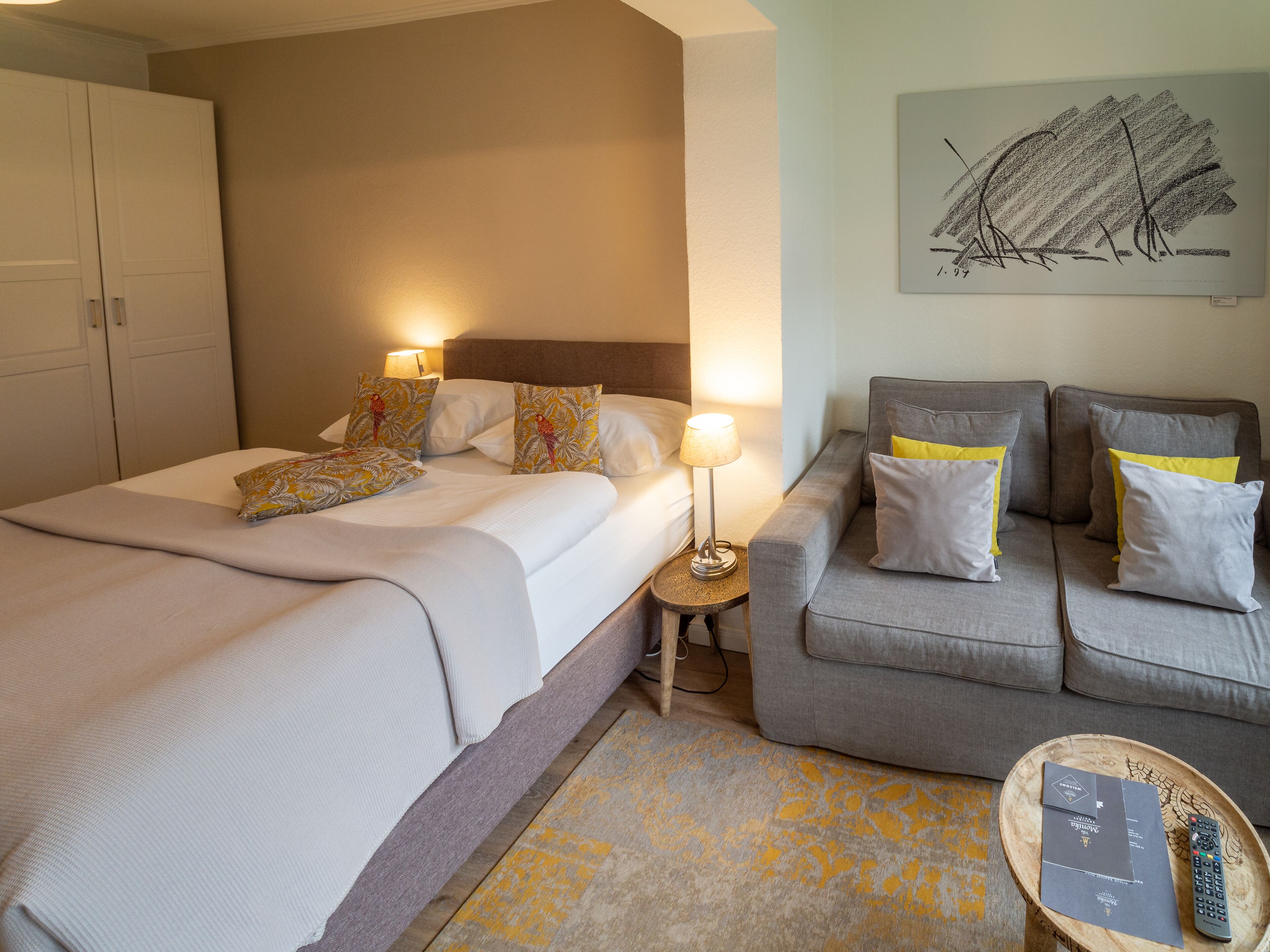 Habitación de hotel con cama, sofá, mesita de noche con lámpara y un cuadro en la pared.