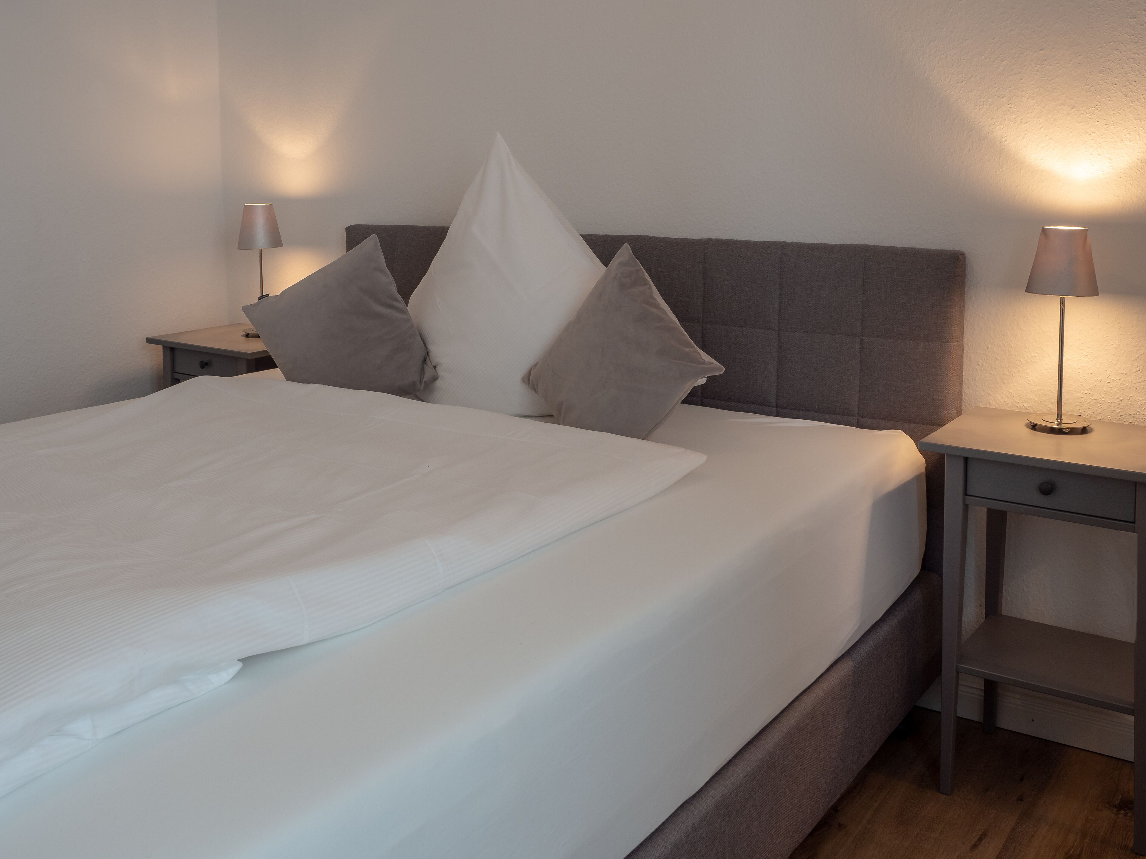 Una cama bien tendida con sábanas blancas y varios almohadones, flanqueada por dos mesitas de noche con lámparas, frente a una pared lisa.