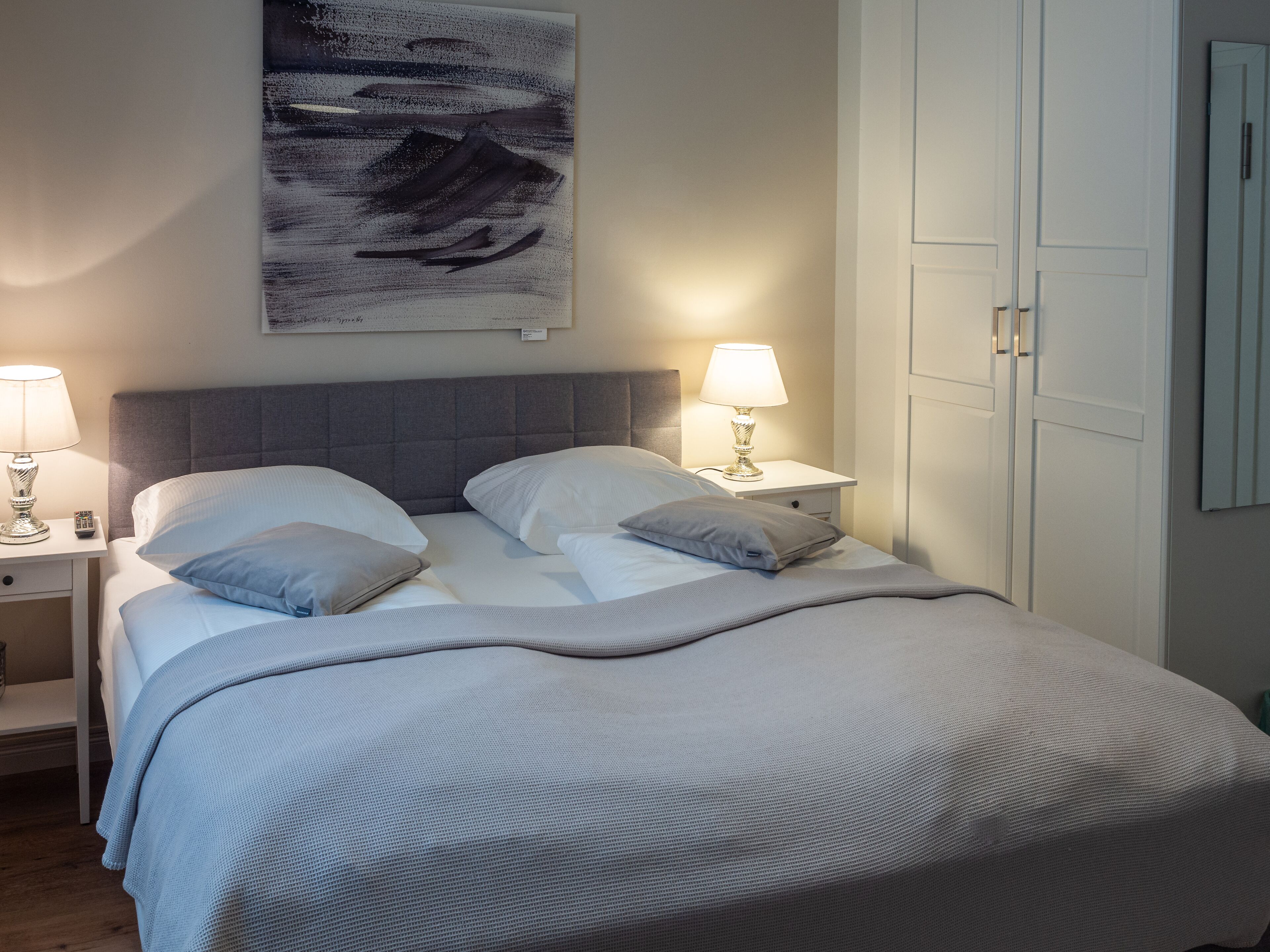 A neatly made bed with a grey headboard, two bedside tables with lamps, and a framed artwork above the bed.