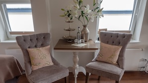 Two upholstered chairs with floral cushions, a wooden table with a vase of white flowers, and a view of the sea through large windows. - Boutique Hotel Villa Monika (Sylt)