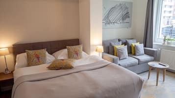 Een hotelkamer met een groot bed, een grijze bank, een klein rond tafeltje en een raam met gordijnen.