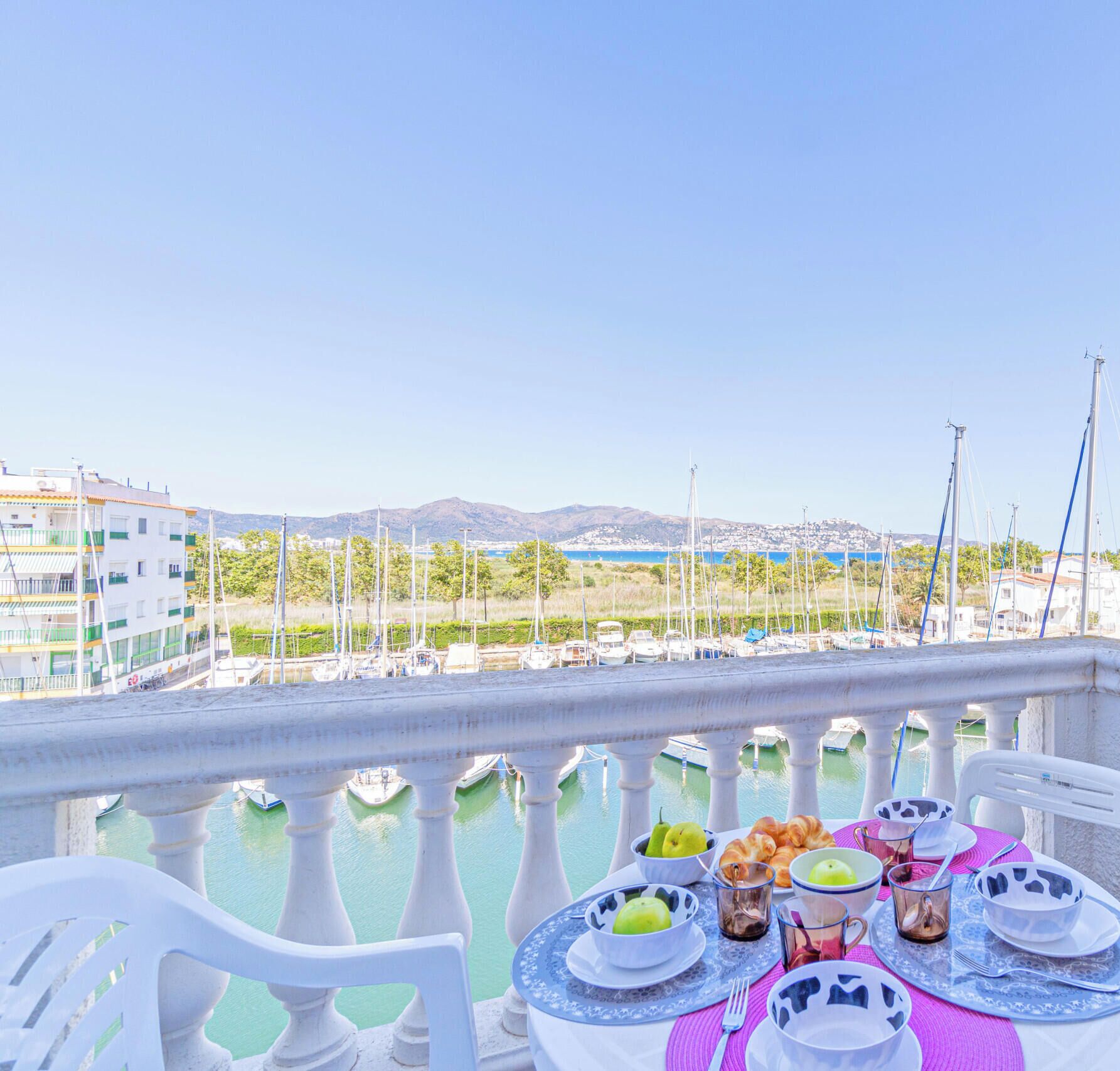 A balcony with a table set for breakfast overlooking a marina with boats and buildings.