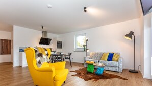 A modern living room with a yellow armchair, a grey sofa with colorful cushions, a dining area with a table and chairs, and a kitchen with a TV and oven. - Apartment 4 Deluxe Apartment 51 sqm, 1-4 persons, balcony and wonderful mountain view (Bayerisch Gmain)