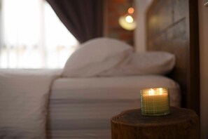 A candle on a wooden stool in a dimly lit room with a bed and a window with curtains.