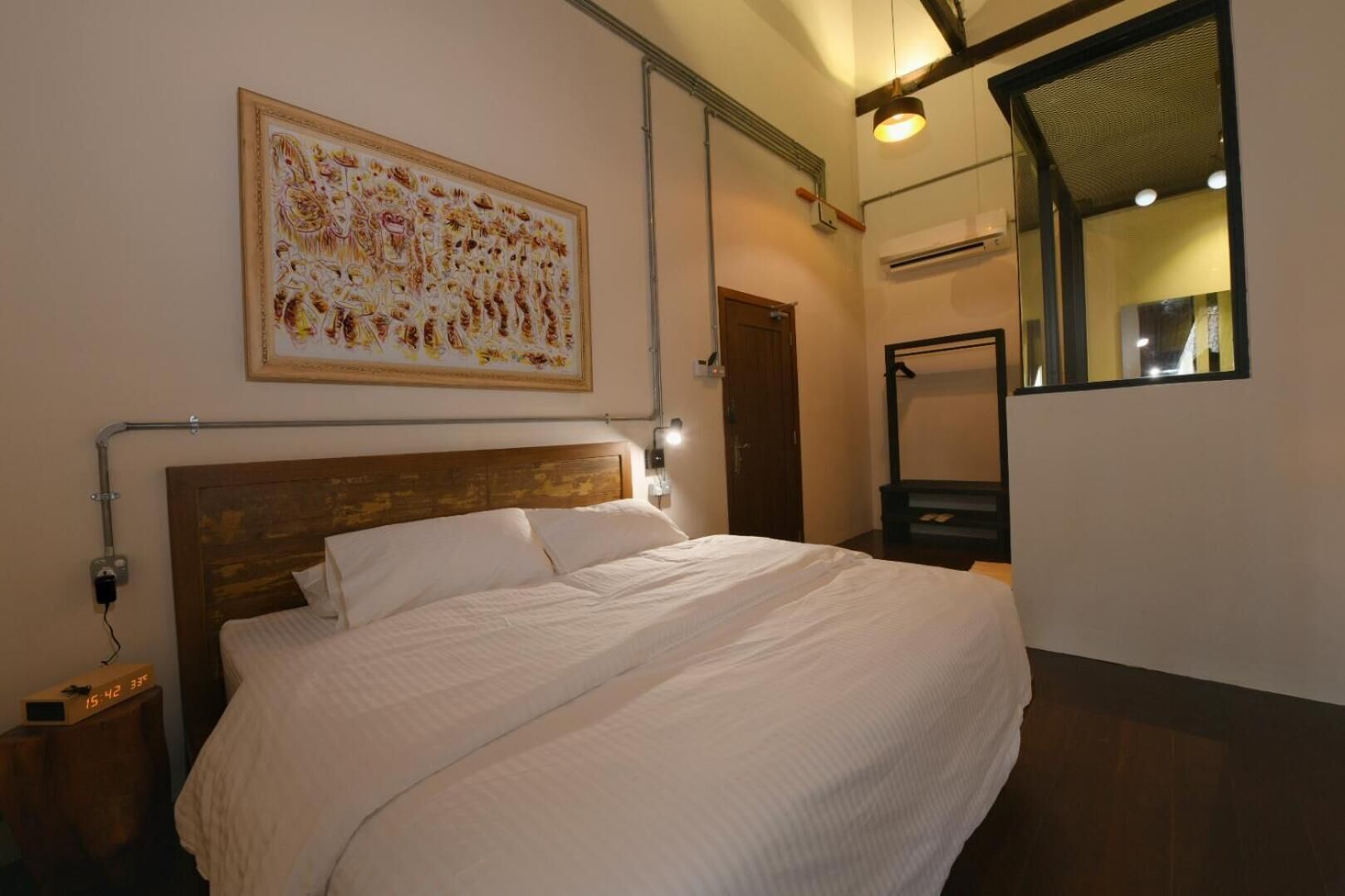 Uma cama bem arrumada num quarto de hotel, com um quadro grande em cima, uma mesa de cabeceira com uma luminária e um espelho na parede.